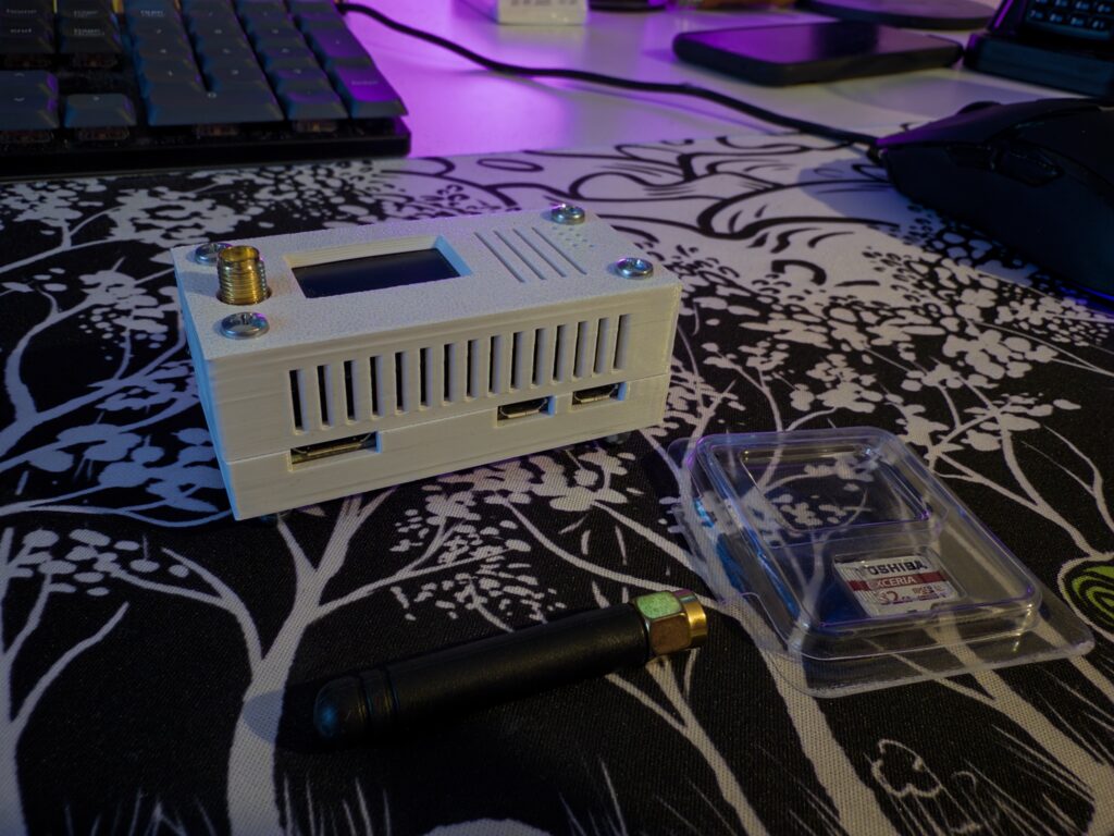 Small box with a screen, ports and an antenna input on a table, accompanied by a small nub antenna and a micro-SD card.